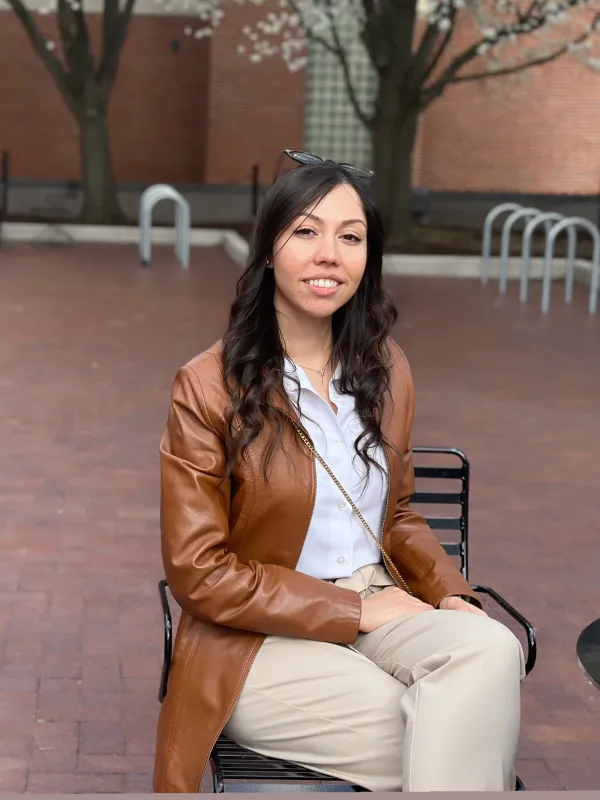 Shima Karimi sitting on a chair wearing a white top, khaki pants, and a brown leather jacket